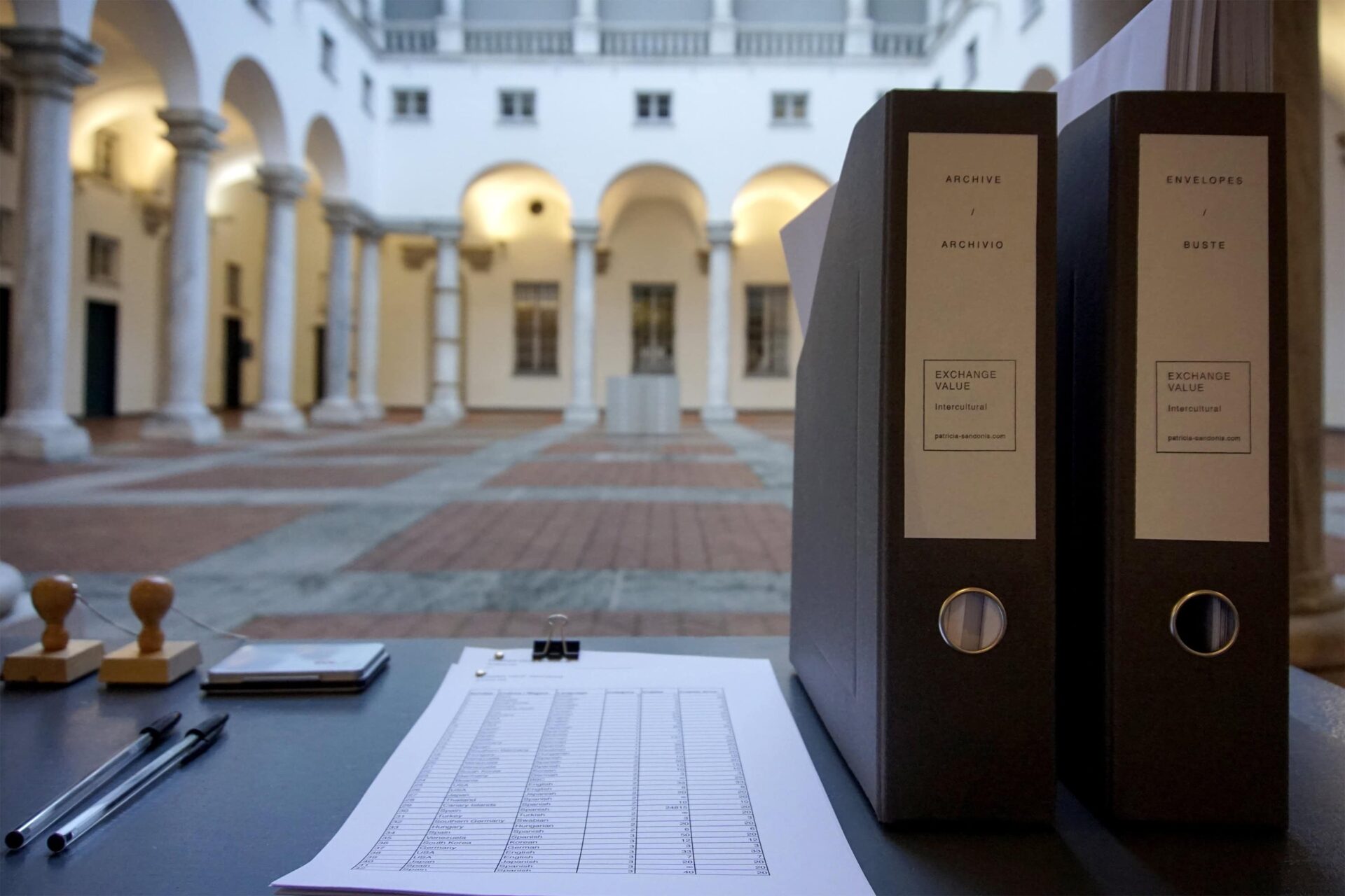 Desk with two black folders standing and a list on paper. The background is a courtyard.