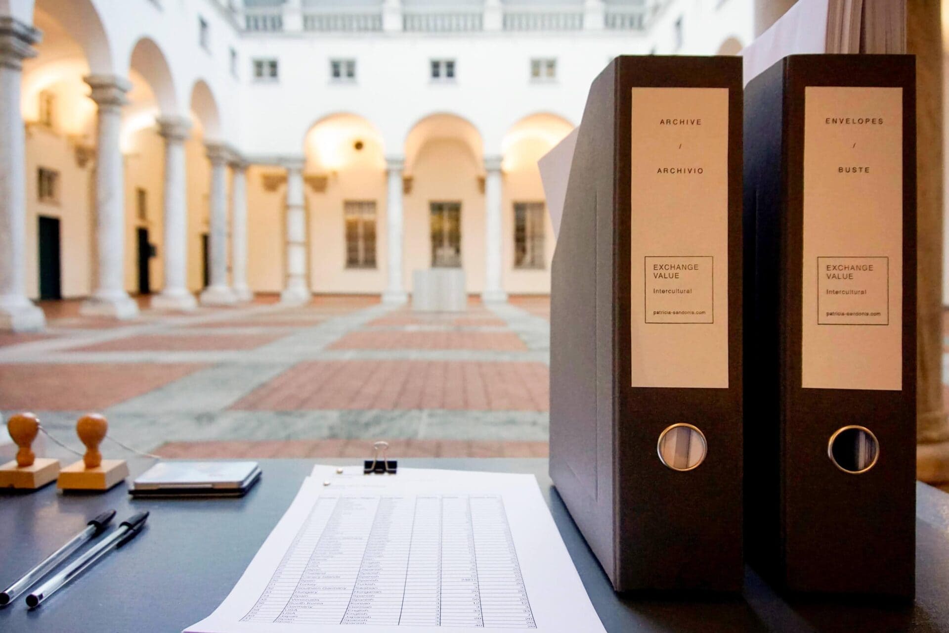 Desk with two black folders standing and a list on paper. The background is a courtyard.