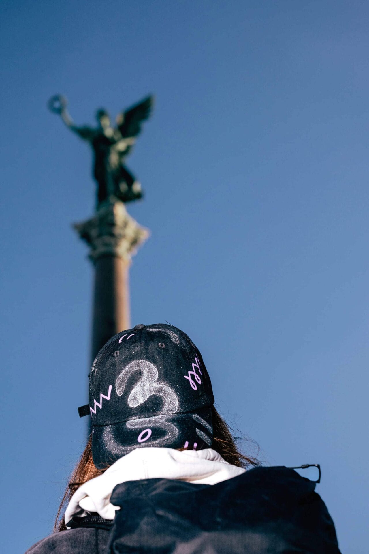 Artist Patricia Sandonis wearing a black cap and looking at a Victory column in Berlin