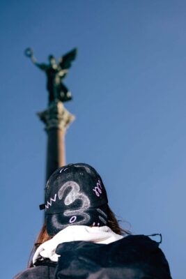 Artist Patricia Sandonis wearing a black cap and looking at a Victory column in Berlin