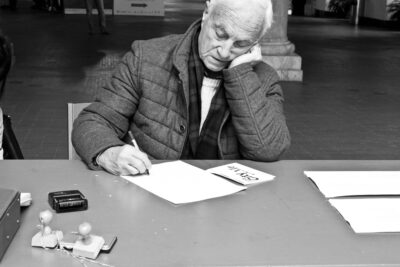 Black and white photography showing an old man sitting at a desk and writing on paper at the art installation Exchange Value Intercultural by Patricia Sandonis