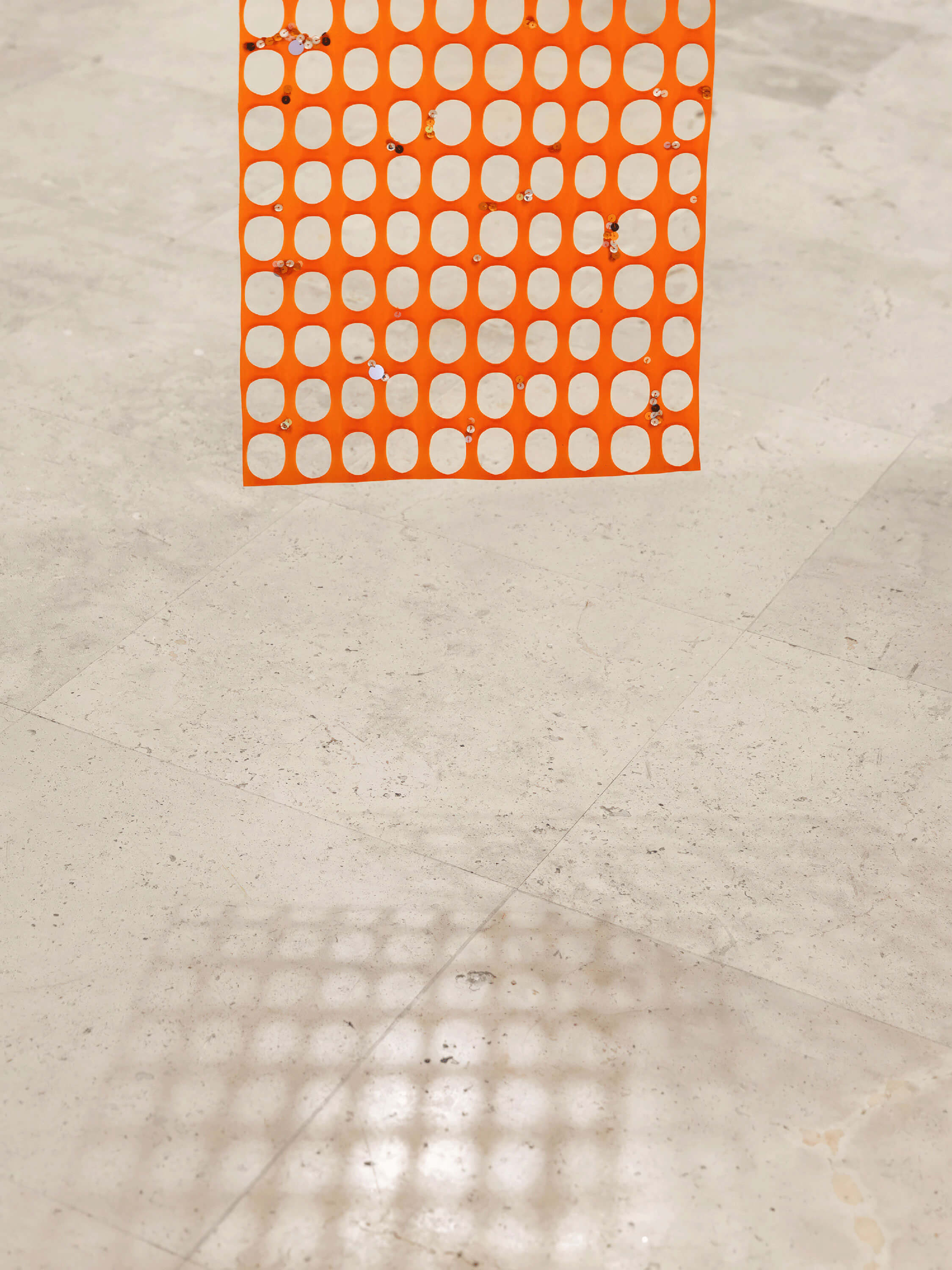 Orange security mesh with sequins hanging on and projecting a shadow on the floor